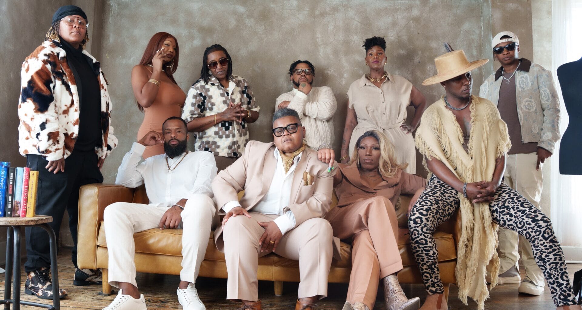 Group of stylish adults posing together in a modern living room, seated and standing along a tan sofa, wearing bold outfits and accessories.