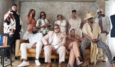 Group of stylish adults posing together in a modern living room, seated and standing along a tan sofa, wearing bold outfits and accessories.