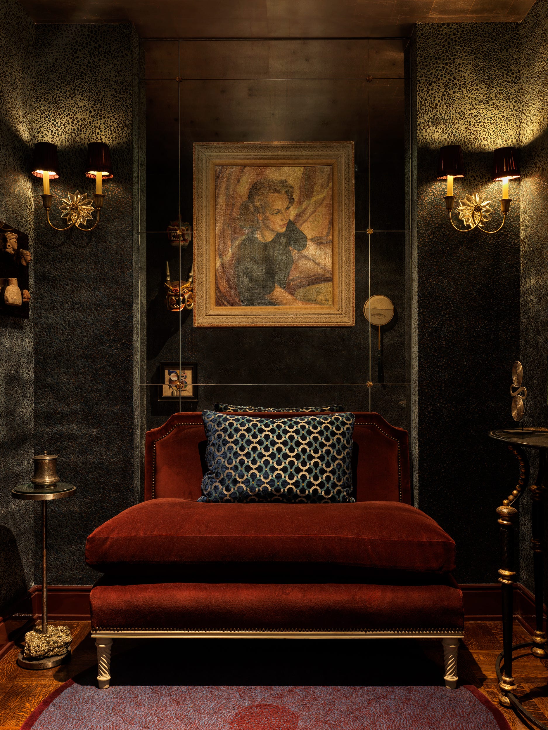 Cozy vintage room with dark walls, elegant sconce lighting, red velvet bench, and a framed portrait.