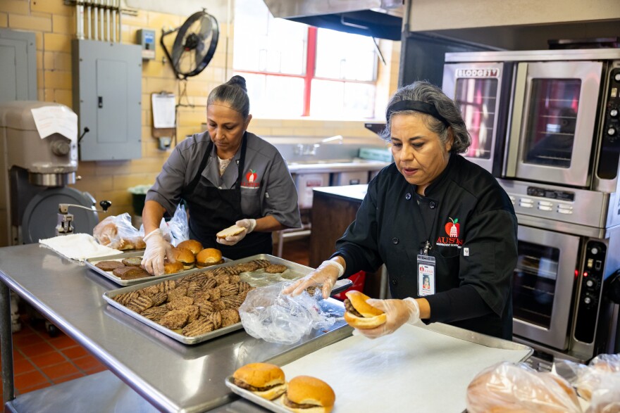 Two women work in a kitchen in a school cafeteria putting together hamburgers.