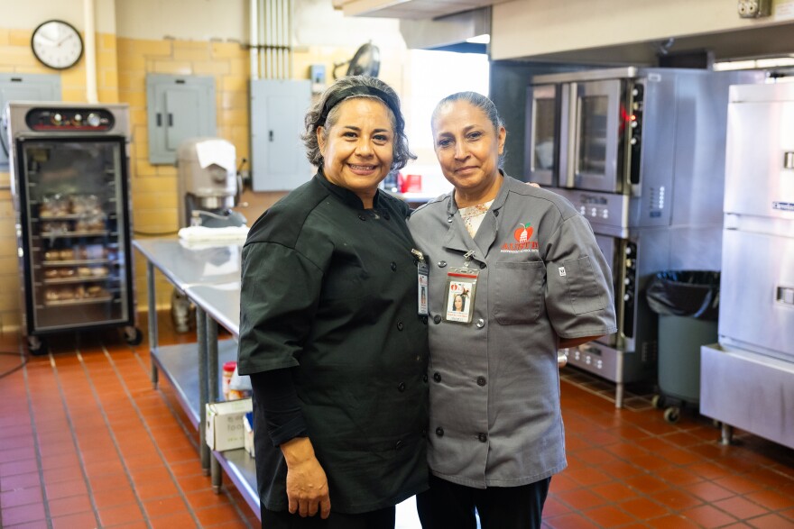 Two women, both wearing chef jackets, pose for a photo together inside a school kitchen.