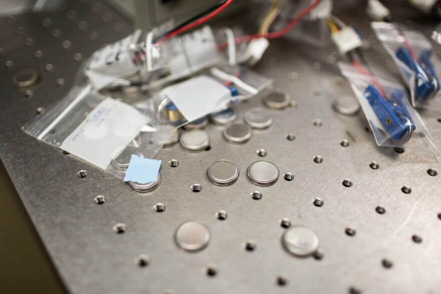 Small battery cells are spread across a metal table with holes in it.