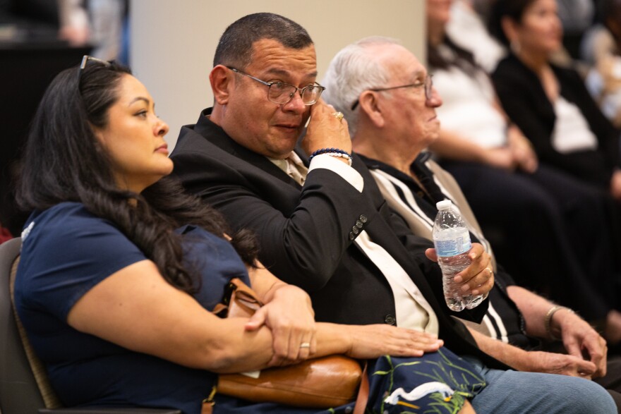 A photo of three people sitting in a courtroom. The person in the center is crying and wiping his tears. 