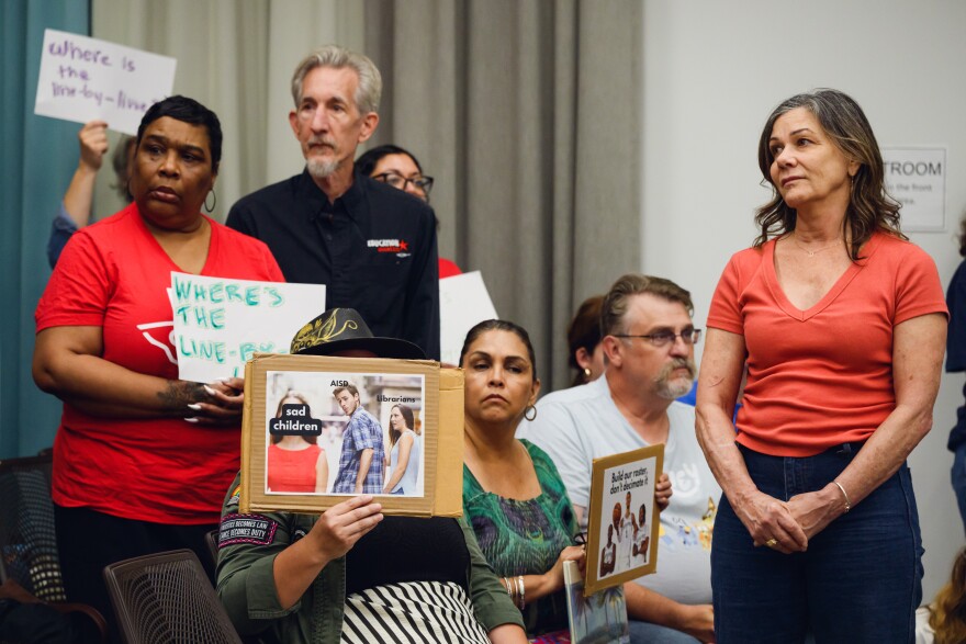A crowd stands together, some holding signs, at a school board meeting.