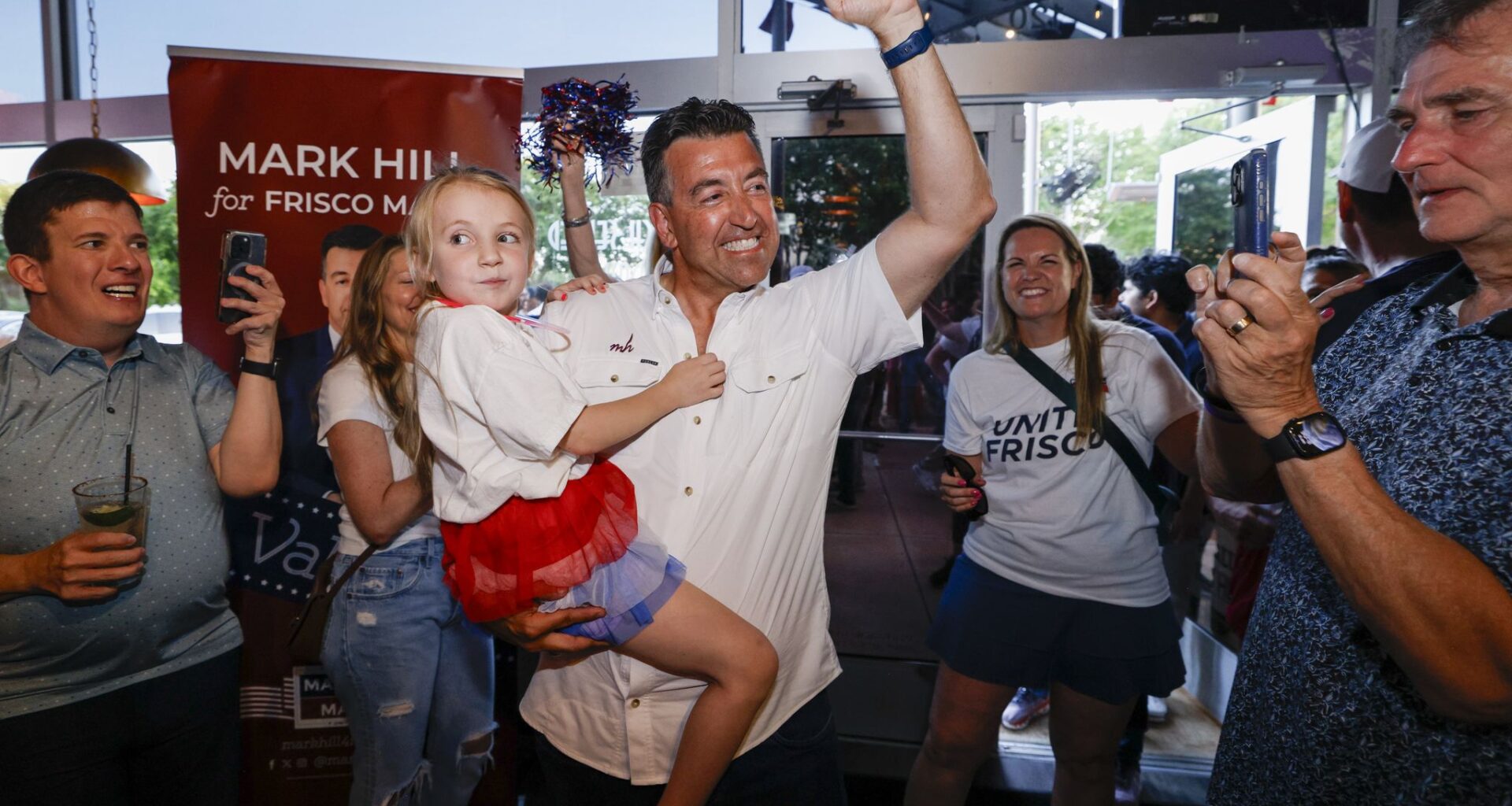 Frisco mayoral candidate Mark Hill pumps his fist as he holds his daughter Emily, 7, as he greets supporters with his wife Tiffany Hill (back right) during a runoff election night watch party at The Frisco Bar &amp; Grill on Saturday, June 13, 2026, in Frisco.