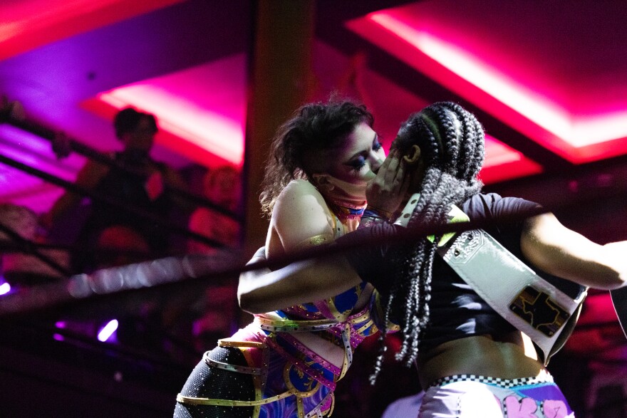 Two competing wrestlers wearing colorful uniforms kiss in the wrestling ring.