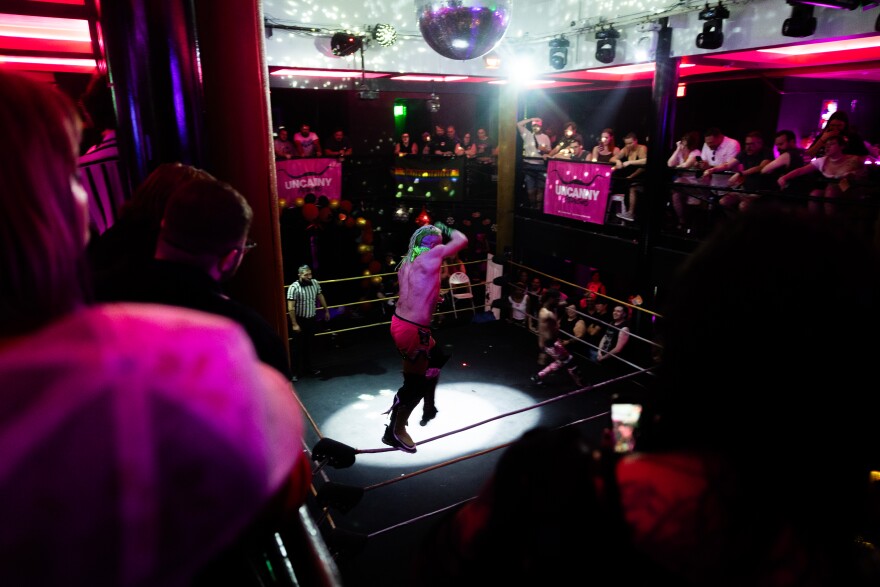 A topdown view shows a wrestling ring with a wrestler jumping off the ropes in the middle of a crowded, tight room with colorful lighting and a mirror ball on the ceiling.