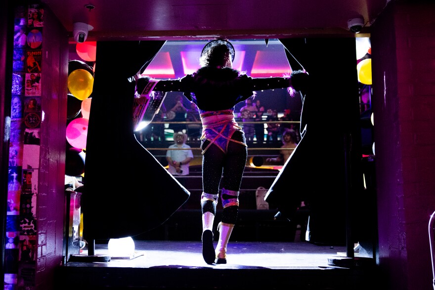 A backlit wrestler throws aside curtains to enter a wrestling arena.