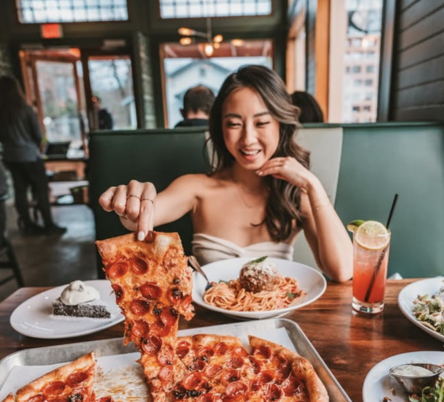 A woman sitting at a table with several dishes of food in front of her. She is holding up a slice of pepperoni pizza.