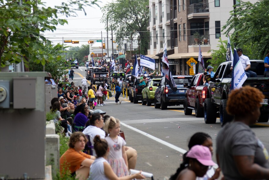 A line of vehicles in a parade formation with flags and banners move down a street lined with spectators.