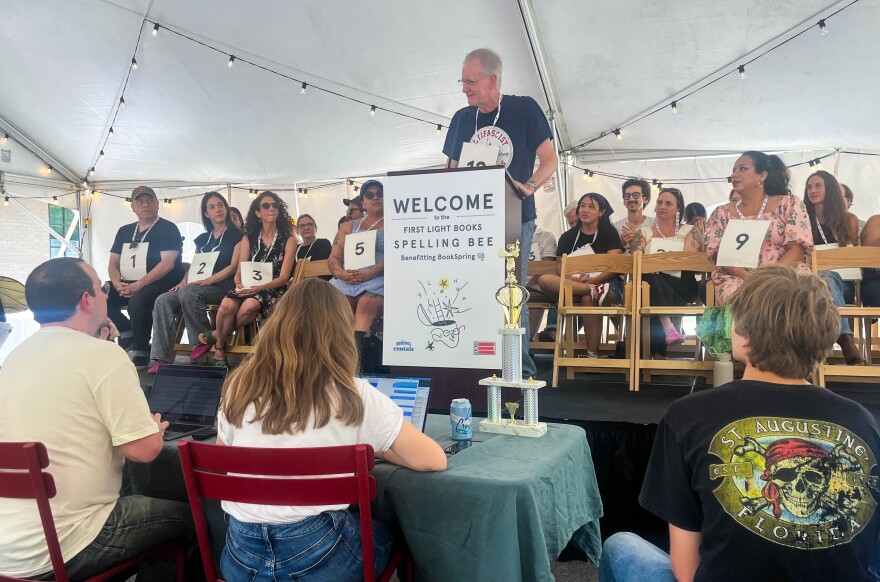 A man stands at a podium on a stage pull of people competing in a spelling bee.
