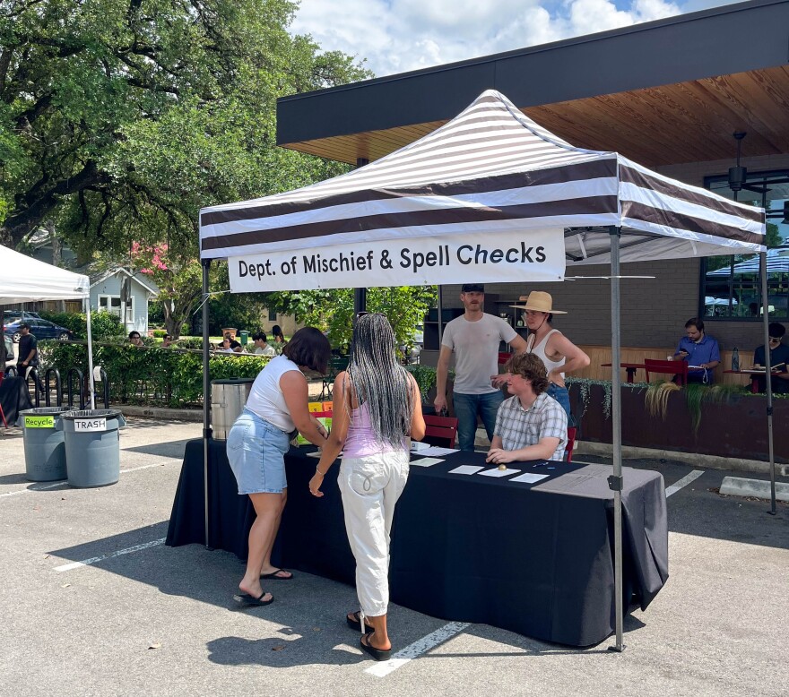 People gather around a tent in a parking lot. It has a sign on it that says "Dept. of Mischief and Spell Checks."