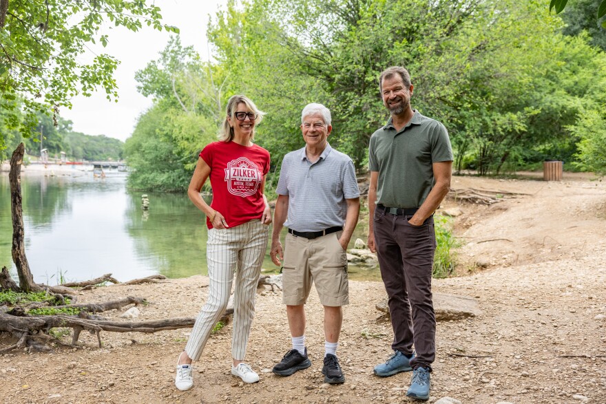 Three people, a woman and two men, pose for a photo at a park near a body of water.