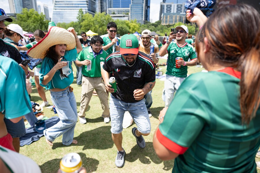 A man in Mexico National Team gear dances while surrounded by other fans at an outdoor watch party.