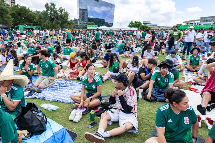People wearing Mexico National Team jerseys sit on blankets across a green lawn. 