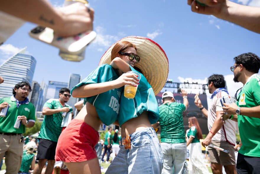 Two women in green Mexico National Team jerseys dance while surrounded by other fans with the city of Austin in the background.