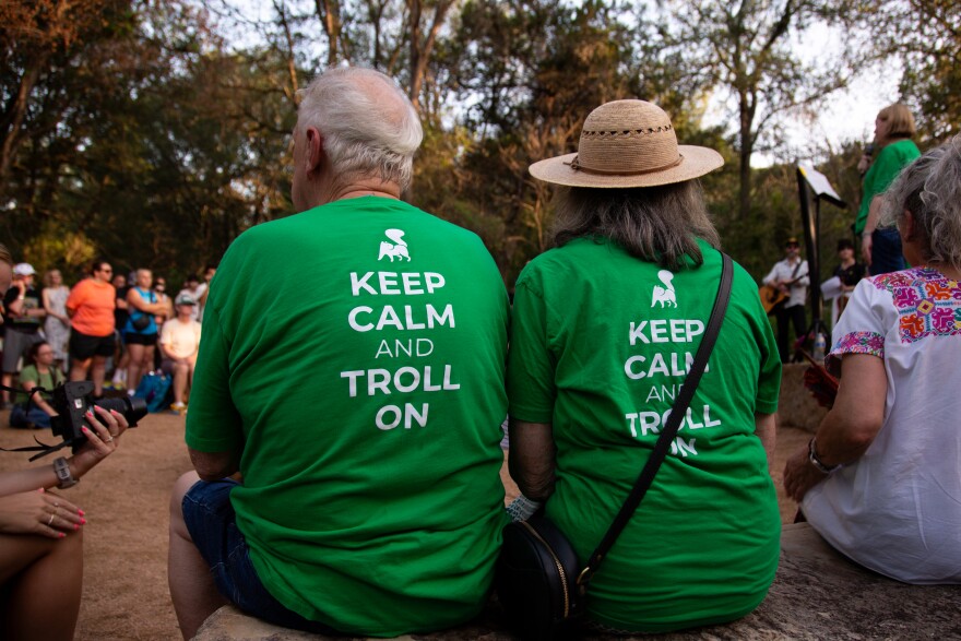 People wear “Keep Calm and Troll On” shirts at a park with other people in the background.