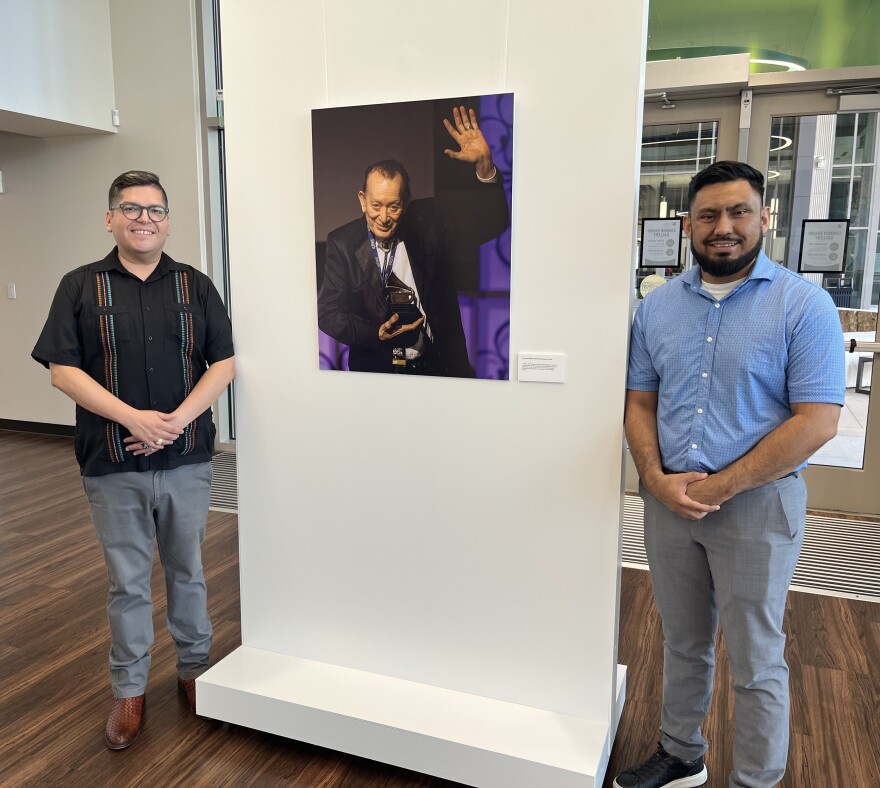 Dr. Eric Castillo, Alamo Colleges Associate Vice Chancellor for Arts, Culture and Community Impact stands with Domingo J. Figueroa, M.Ed., Program Manager of Arts, Culture, & Community Impact for the ACD. Between them is an enlarged photograph of Flaco Jimenez receiving the Grammy Lifetime Achievement Award in 2015.