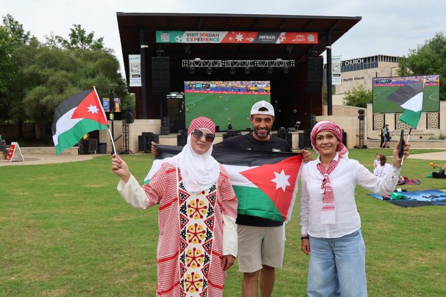 Three people dressed in traditional Jordanian attire pose outdoors holding Jordan flags.