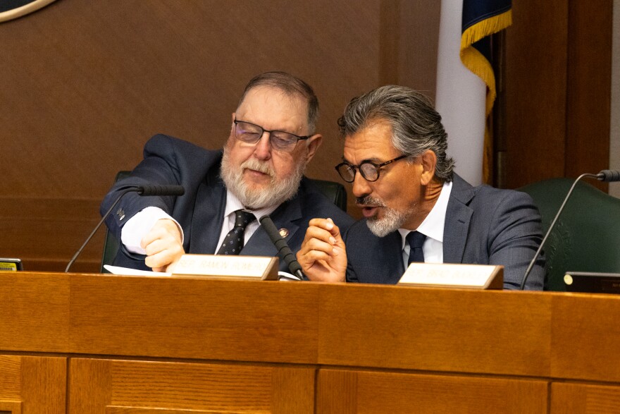 Two men sitting at a dais at a public hearing lean in and speak to each other. One man points to a sheet of paper.