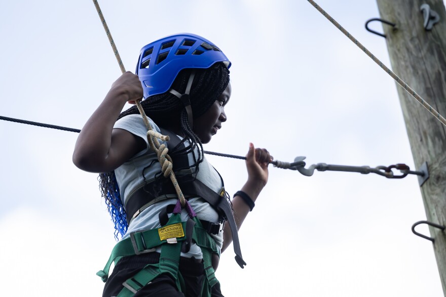 A young teenage girl navigates a high ropes course with a helmet and harness strapped on.