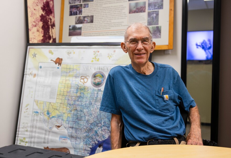 A man poses for a photo wearing a blue shirt and sitting at a table in front of a map of Texas.
