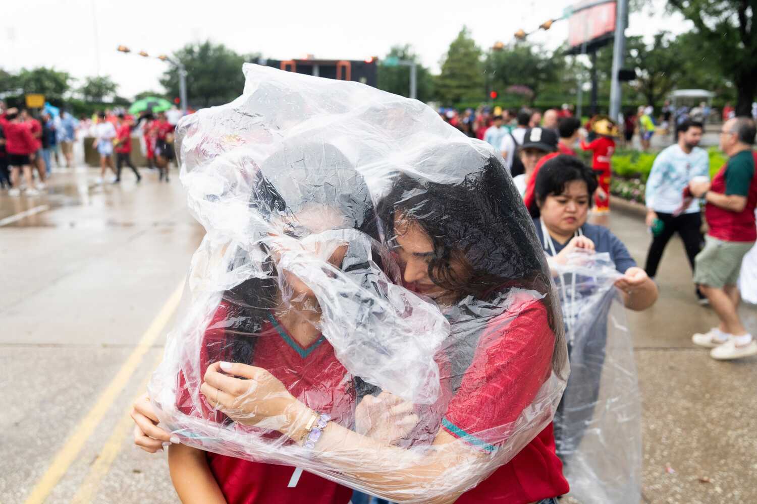 Two women in red soccer jerseys drape a single poncho over their heads as rain falls. 