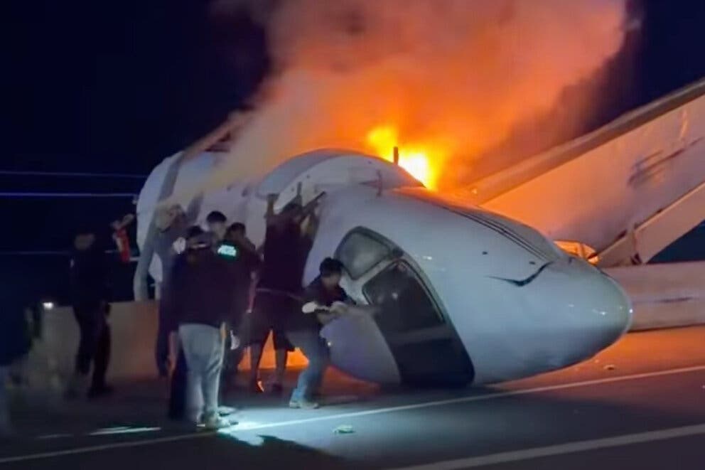 The burning wreckage of a small passenger aircraft, with its nose section resting on its side on a highway. Some people are trying to open the door and break through its windshield.