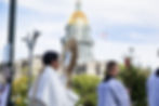 Catholic procession with priests carrying a gold monstrance and red candle before a domed capitol building under a bright sky.