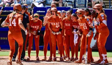 Female softball players stand together celebrating another player coming towards them on a softball field.