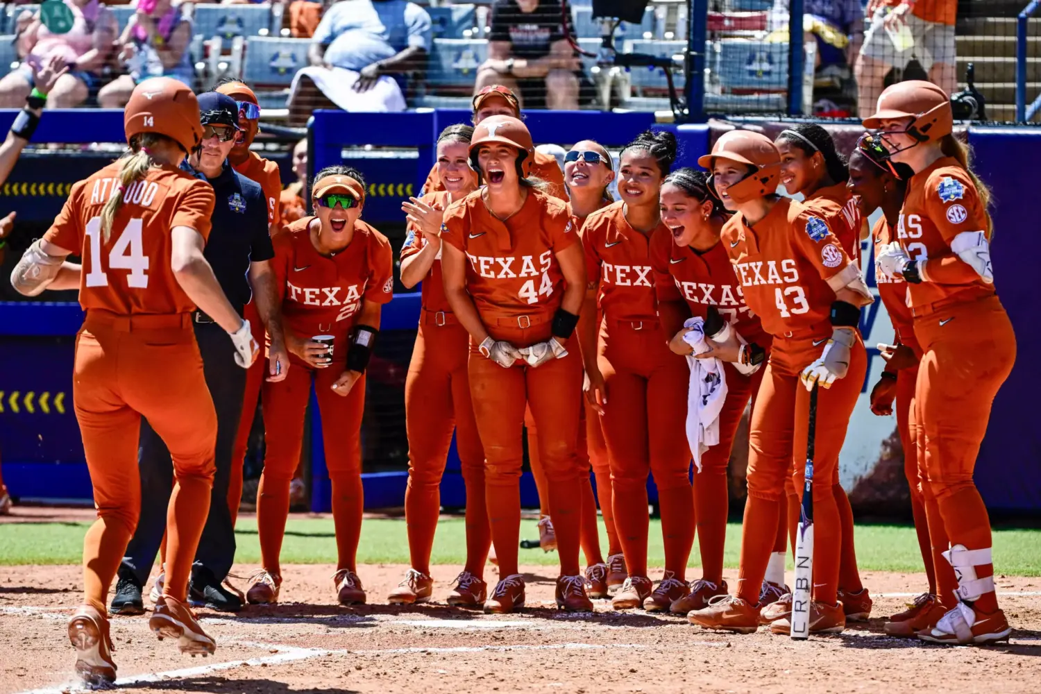 Female softball players stand together celebrating another player coming towards them on a softball field.