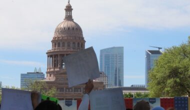 People, whose faces are not shown, hold up signs with the Texas Capitol in the background.