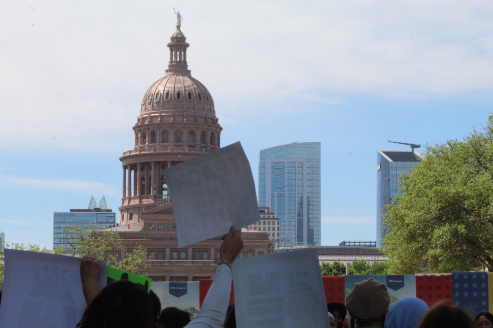 People, whose faces are not shown, hold up signs with the Texas Capitol in the background.