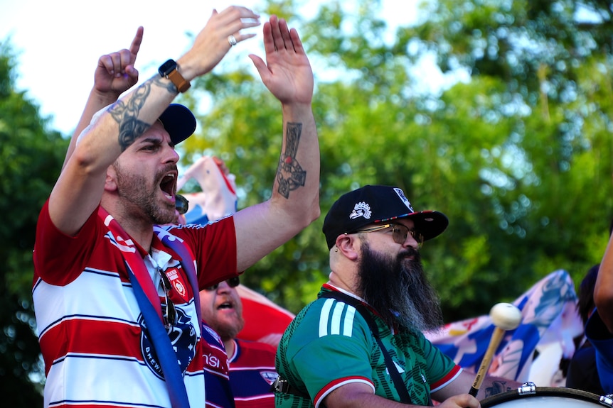 A man wearing a red and white FC Dallas supporter shirt yells and claps his hands in the air.