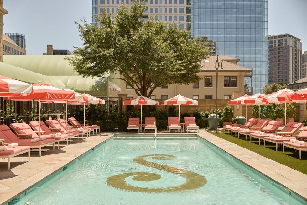 Outdoor pool with pink loungers and red umbrellas, surrounded by buildings and a large tree.