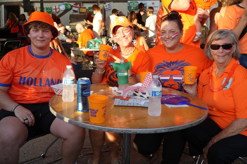 Four people wearing Orange Netherlands supporter shirts smile while seated around a table.