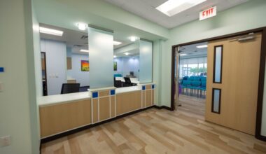 A modern medical office reception area with light green walls, wooden flooring, and a counter with privacy screens. An open door leads to a waiting room with teal chairs.