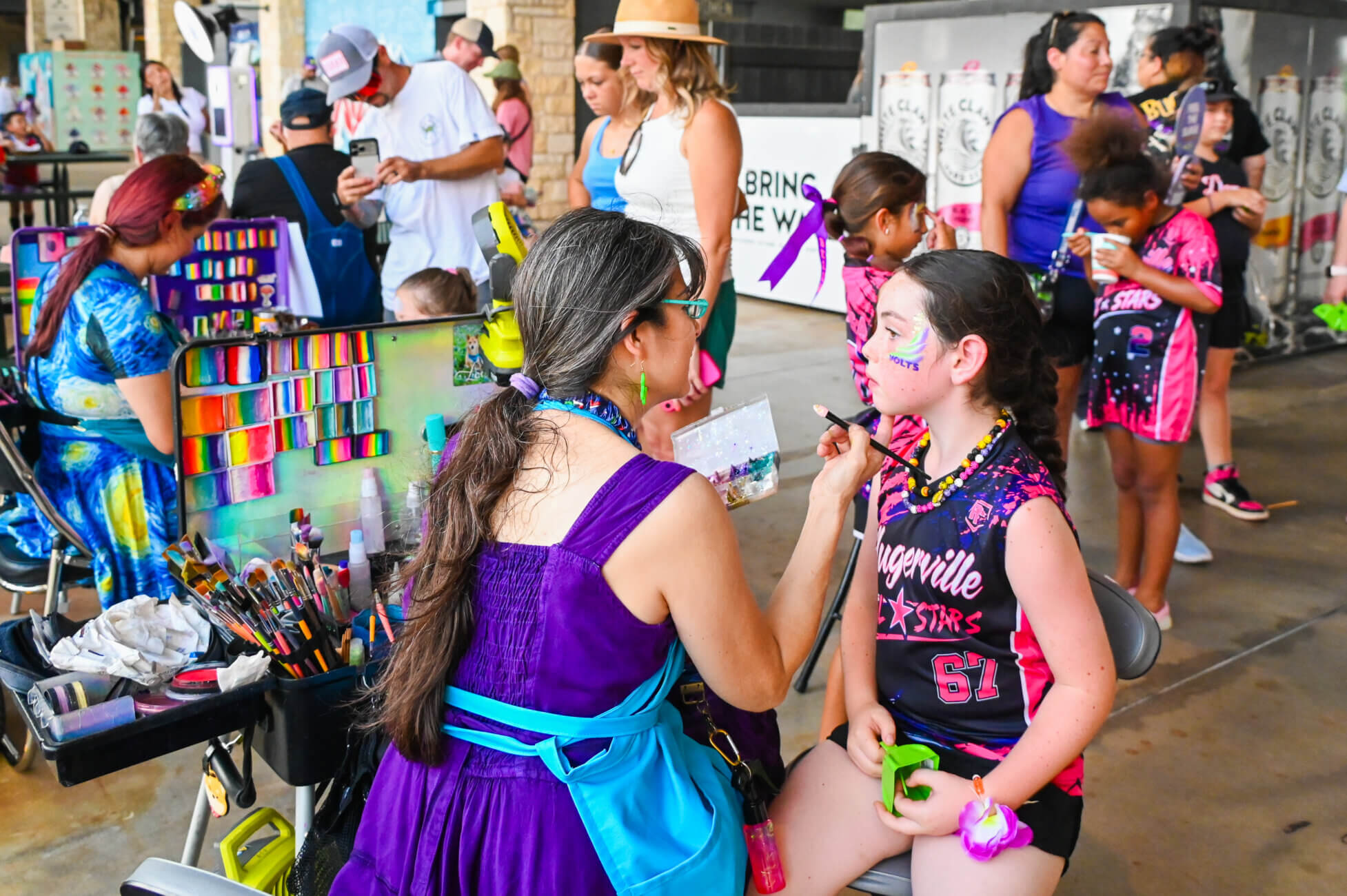 A woman in a purple dress paints a young girls face at a colorful, lively event with other children and adults participating in activities in the background.