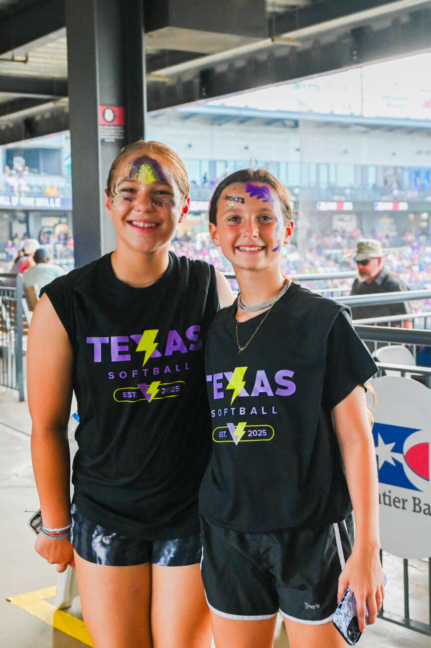 Two smiling girls wearing matching Texas Softball shirts and face paint pose together inside a sports stadium, with crowds and stadium seating visible in the background.
