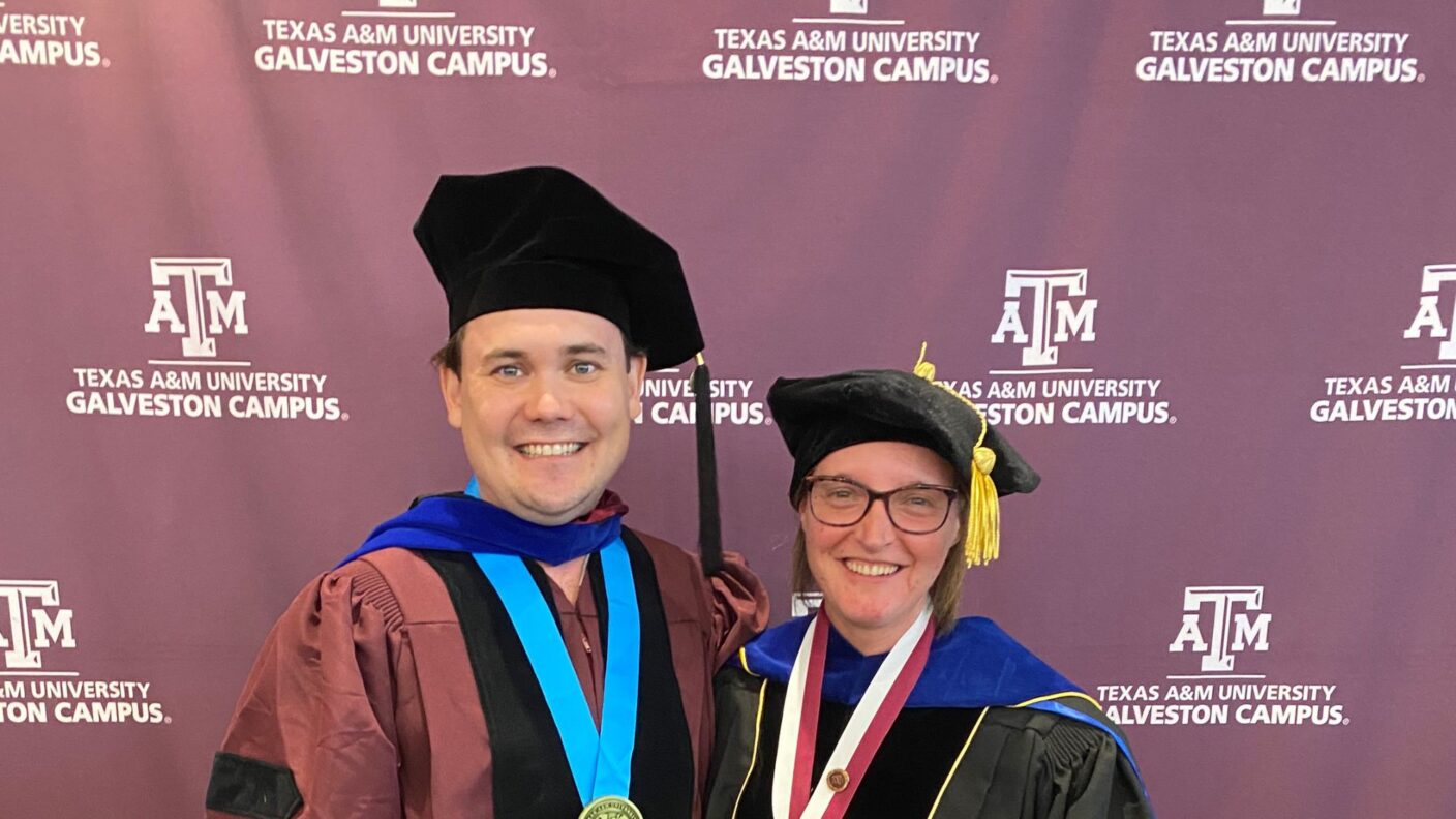 Two scientists pose in their caps and gowns in front of a Texas A&M University at Galveston backdrop.