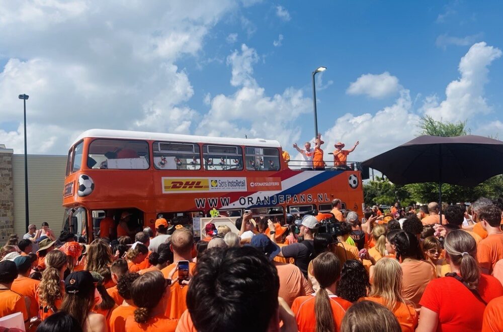 A sea of people in orange t-shirts are crowded around a large, orange double decker bus