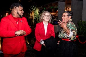 Three adults laugh and chat at a bar: two wearing bright red jackets and one wearing a camo-pattern jacket with glasses, indoors at night.