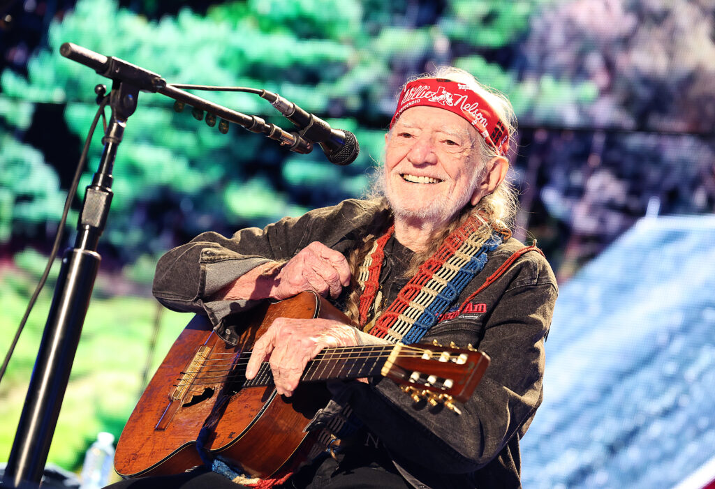 Willie Nelson is seen holding his guitar on stage with his signature bandana tied around his head.