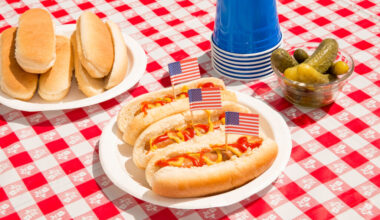 America and hot dogs. Fourth of July concept for carefree summer BBQ, unhealthy eating of junk food, Independence Day or Memorial Day backyard BBQ. Picnic food, no people. July 4th foods featured are hot dogs with ketchup and mustard and pickles. There are patriotic American flag toothpicks as decoration in the festive food. Paper plates and disposable plastic cups are also on the checkered tablecloth. Shot during sunny day outdoors on vacation. Part of a series.