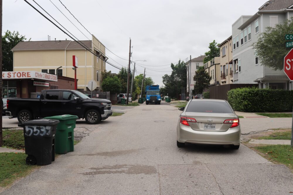 A car reverses on a street in Houston's Cottage Grove neighborhood, blocked by a garbage truck on the narrow road.