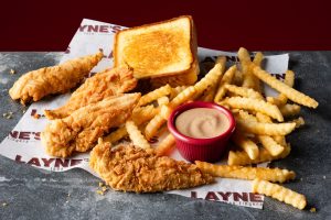 Basket of fried chicken tenders, crinkle-cut fries, and a small cup of dipping sauce on branded parchment mustard-yellow crust bread slice in the background