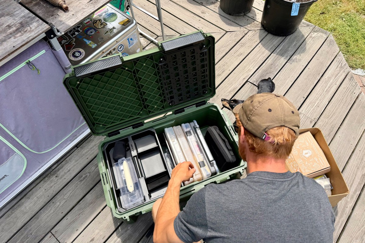 Man organizing tackle boxes inside an open green Plano box on a deck