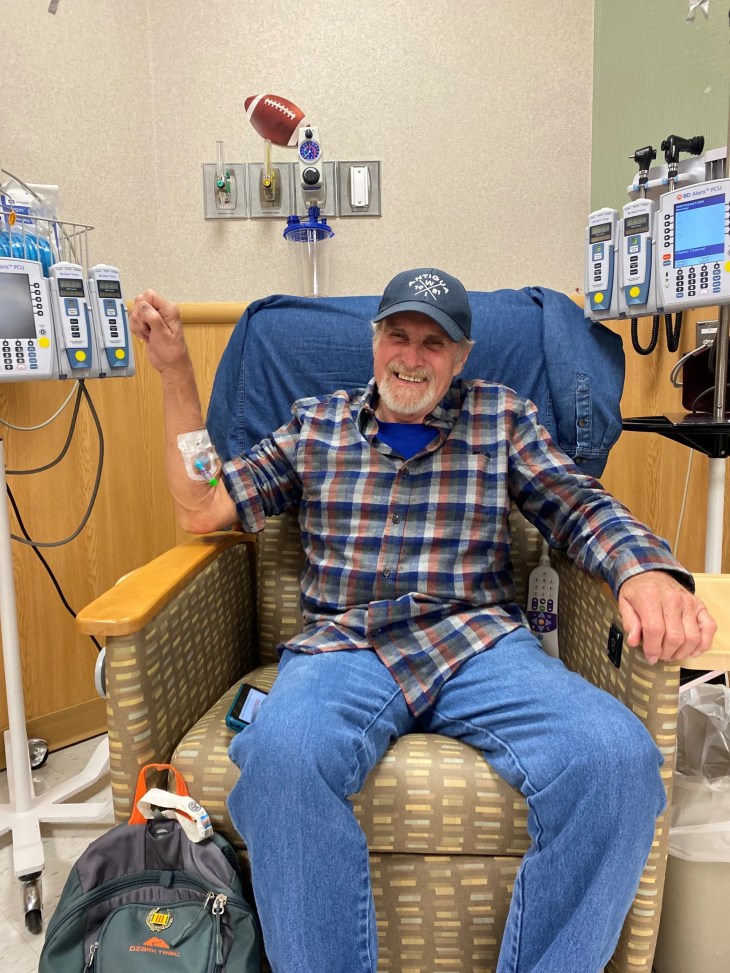 A person sits in a medical chair with an IV in one arm, raising the other arm in a gesture of strength. Medical infusion pumps and equipment surround the chair, and a bag rests on the floor nearby.