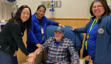 A person sits in a medical chair receiving an IV while three healthcare staff members stand nearby with hands resting on the patient’s arm and chair, representing support during cancer treatment.