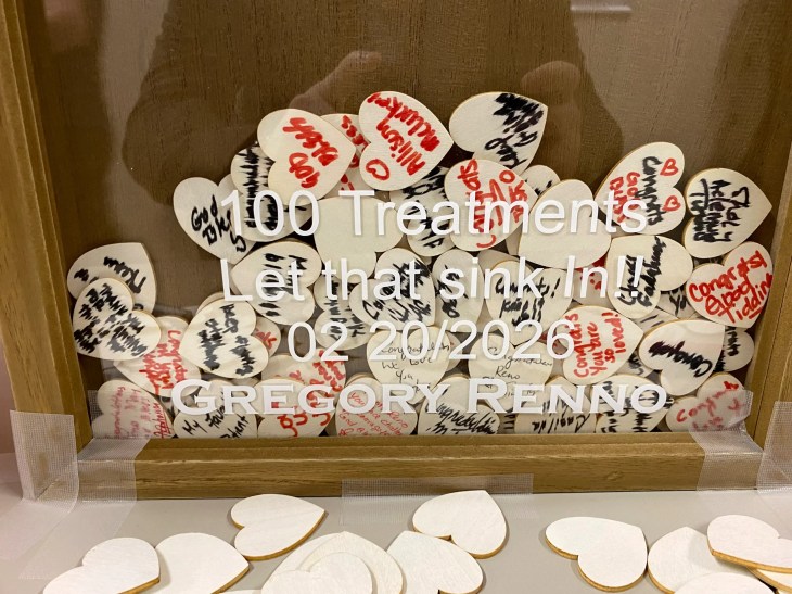 A display box filled with small wooden heart shapes, each with handwritten messages, sits on a surface. Text on the front reads “100 Treatments, Let that sink in!! 02/20/2026 GREGORY RENNO,” with additional blank heart shapes placed outside the box.